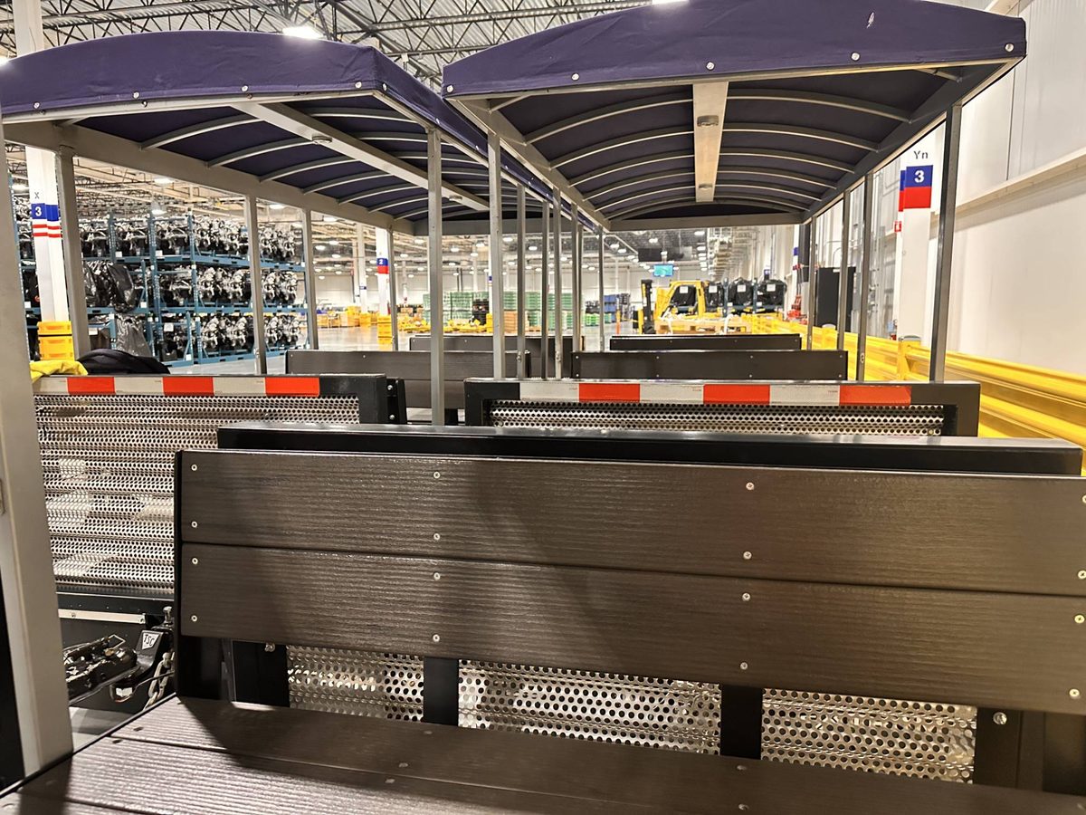 View along a FlexTram train inside an engine assembly plant, with racks of engines and a forklift beyond the canopy