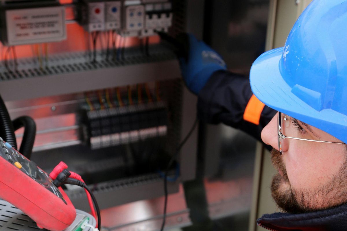 Construction electrician in safety gear and blue hardhat working on an industrial control panel with multimeter and colorful wiring visible