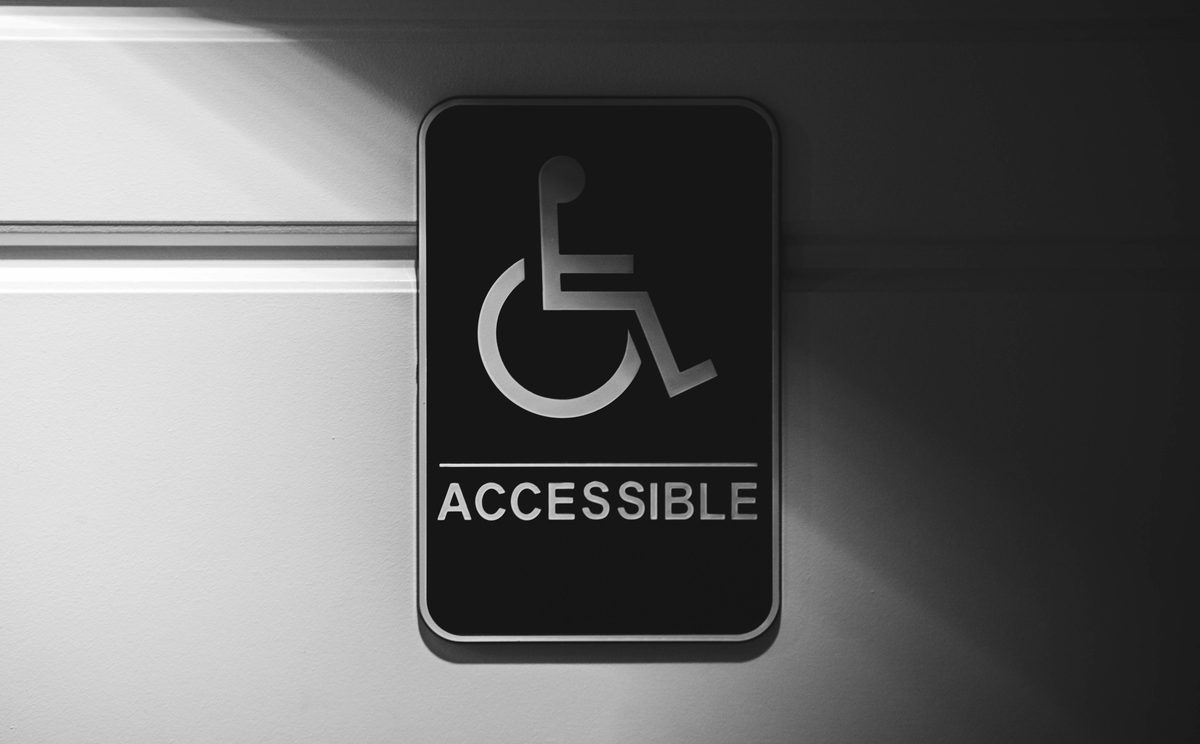 Black-and-white photograph of a black 'ACCESSIBLE' wheelchair sign mounted on a wall, lit dramatically from above