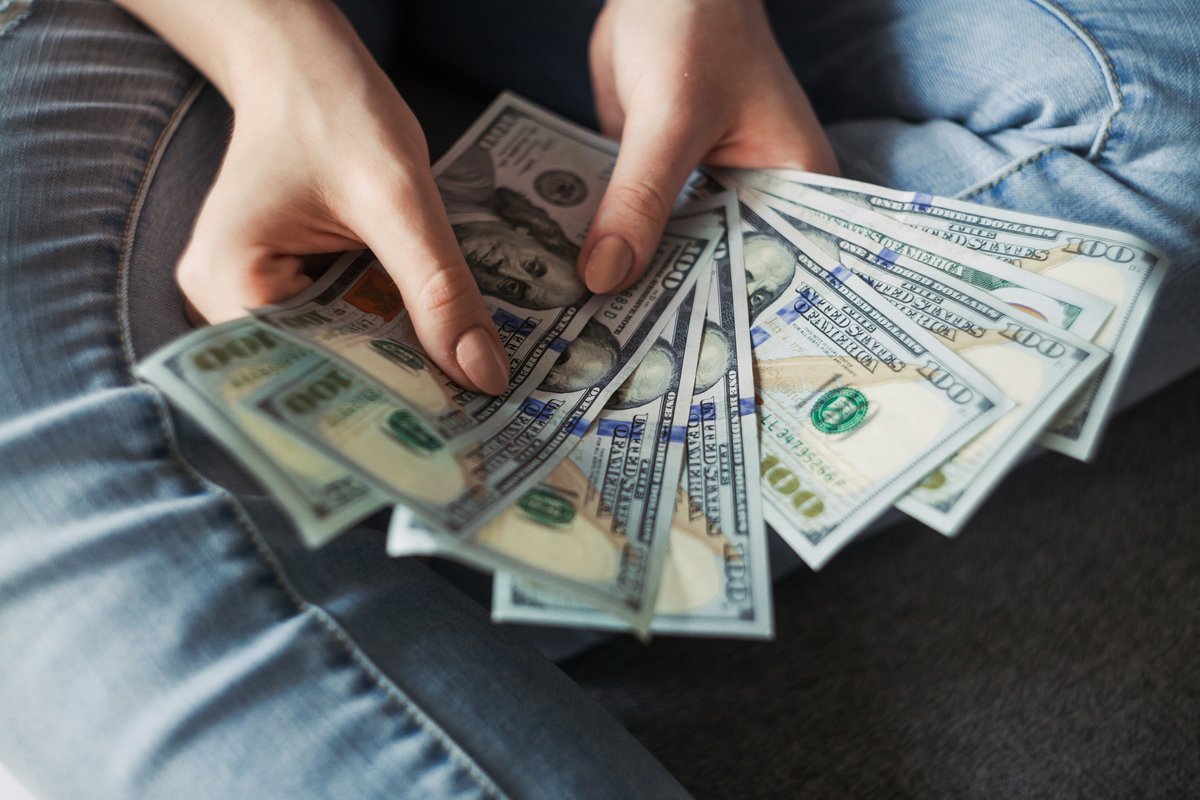 Hands counting a fan of US one-hundred-dollar bills against a denim background, evoking the hidden costs scattered through a venue budget