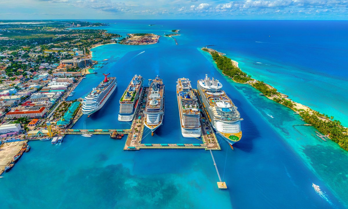 Aerial view of Nassau Bahamas cruise port with five cruise ships docked side-by-side at the central pier — Royal Caribbean, Carnival, Norwegian, and MSC vessels visible — flanked by Paradise Island and the city of Nassau, with turquoise Caribbean water in every direction
