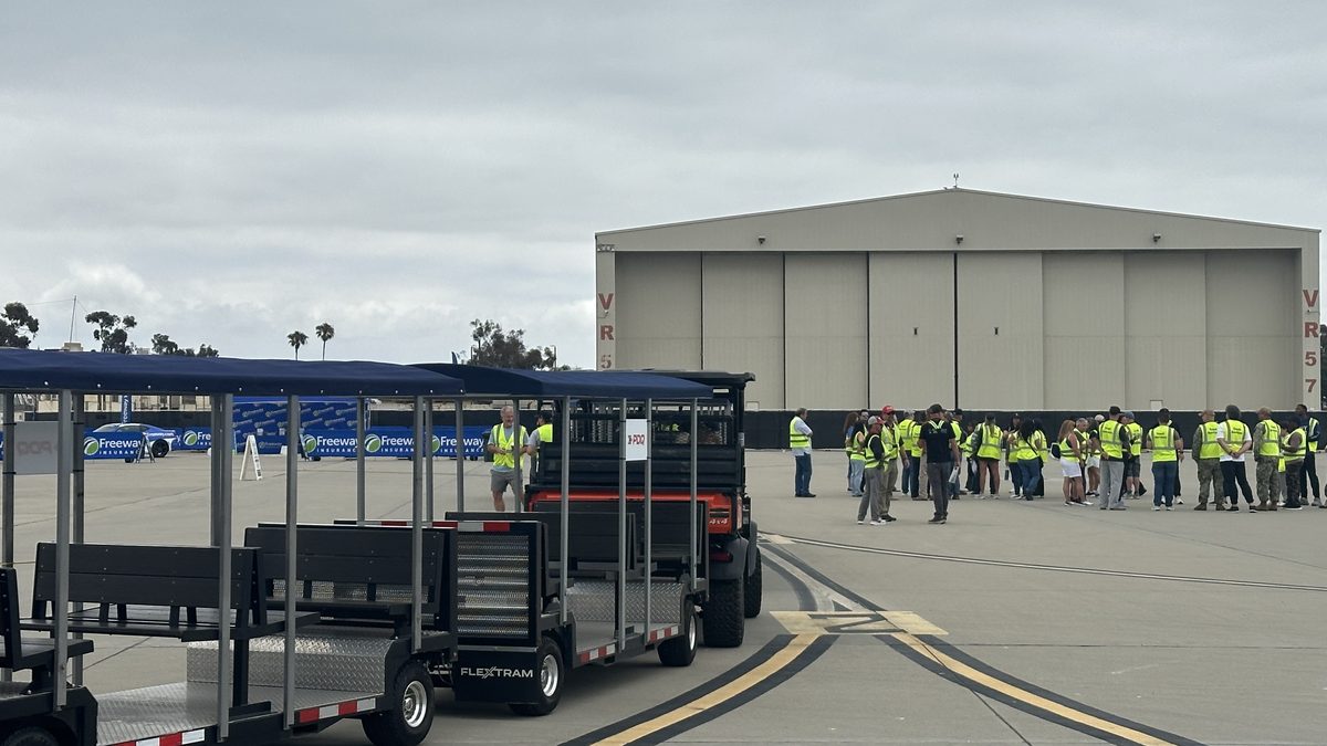 A FlexTram crew shuttle staged on an airfield apron beside a group of event staff in safety vests gathered for transport