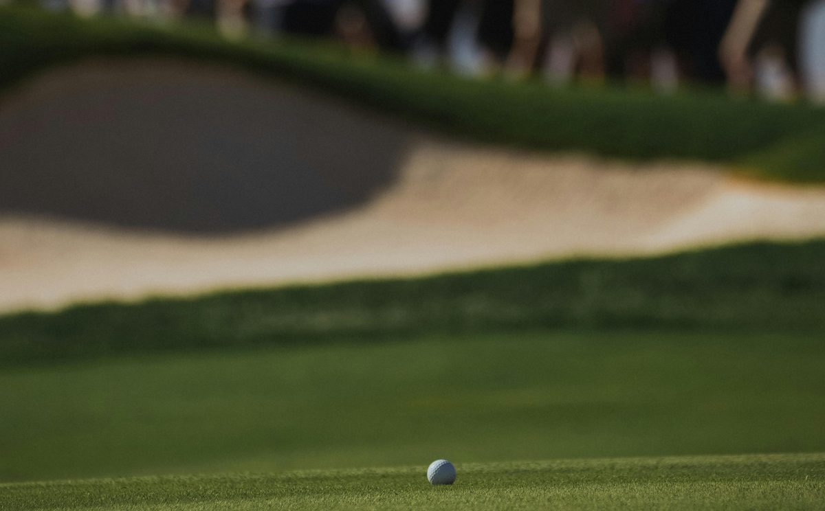 Golf ball at rest on a tournament green with a sand bunker behind it and a crowd of spectators silhouetted in the background