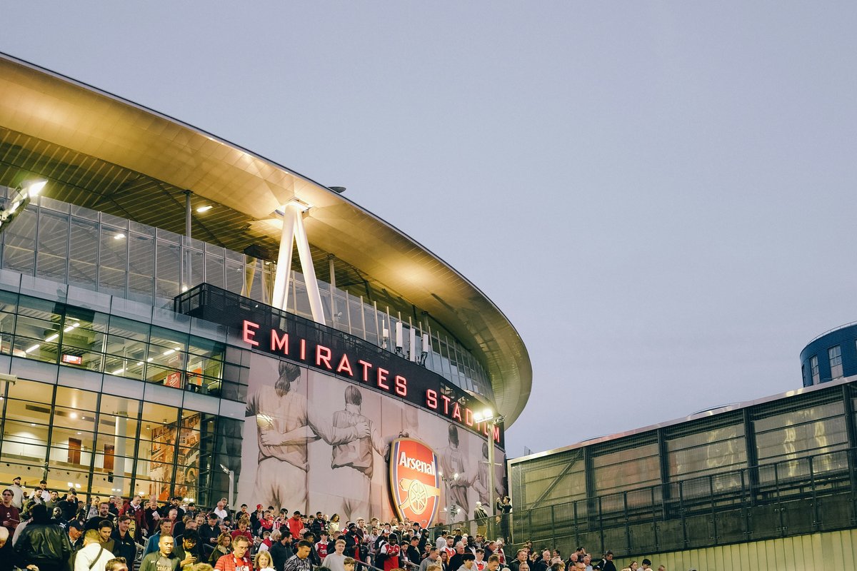 Crowds of fans streaming out of Emirates Stadium at dusk after a match, with the illuminated stadium facade and Arsenal crest visible above the moving crowd