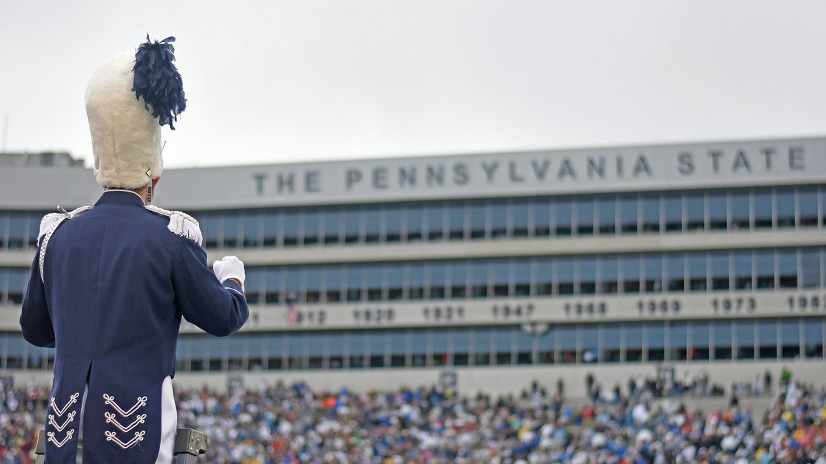 A Penn State drum major in full uniform stands on a conductor's ladder facing a packed Beaver Stadium and the press box reading THE PENNSYLVANIA STATE on college football game day