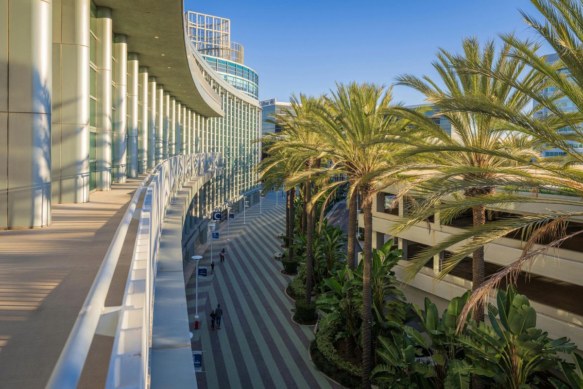 Anaheim Convention Center exterior with curved glass facade, palm-lined pedestrian plaza, parking structure, and adjacent Hilton hotel tower in afternoon light