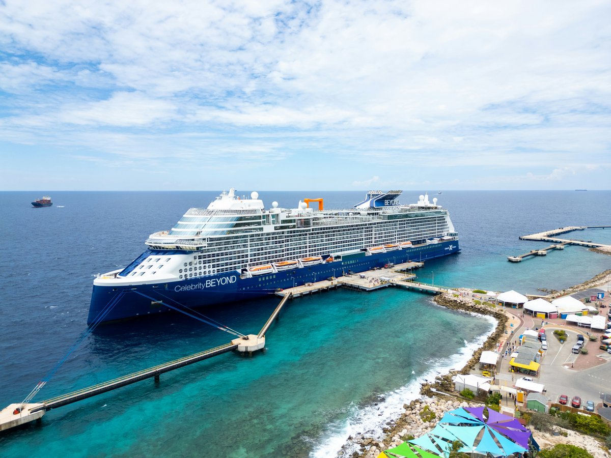 Celebrity Beyond cruise ship docked at a Caribbean port with turquoise water, market stalls, and colorful pavilions visible alongside the pier