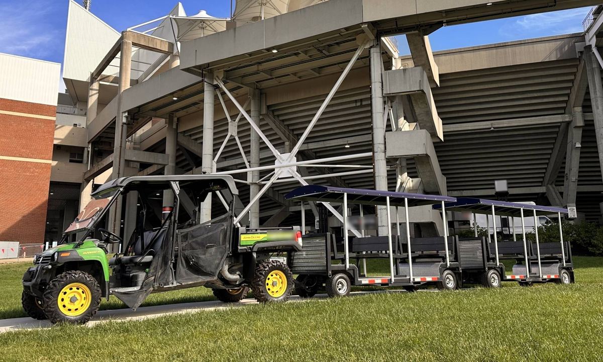 FlexTram tram cars towed by a utility vehicle on the lawn outside a college football stadium