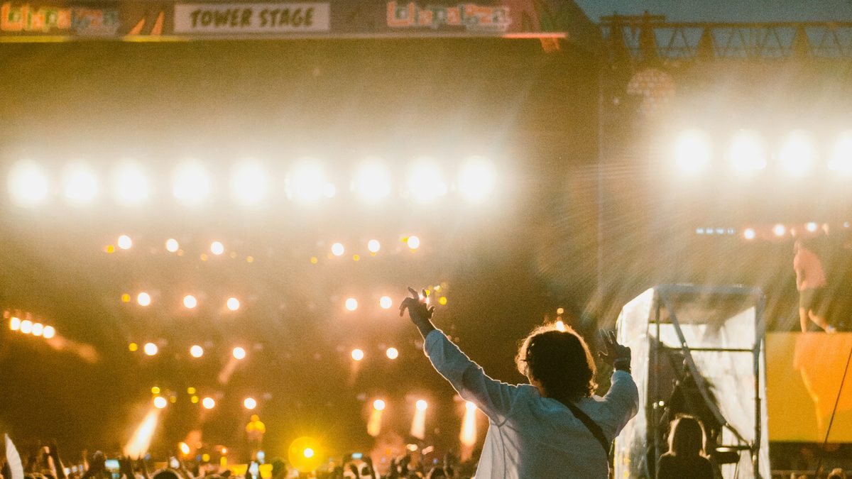 A fan with arms raised in a dense nighttime crowd facing the brightly lit Tower Stage at Lollapalooza in Grant Park, downtown Chicago