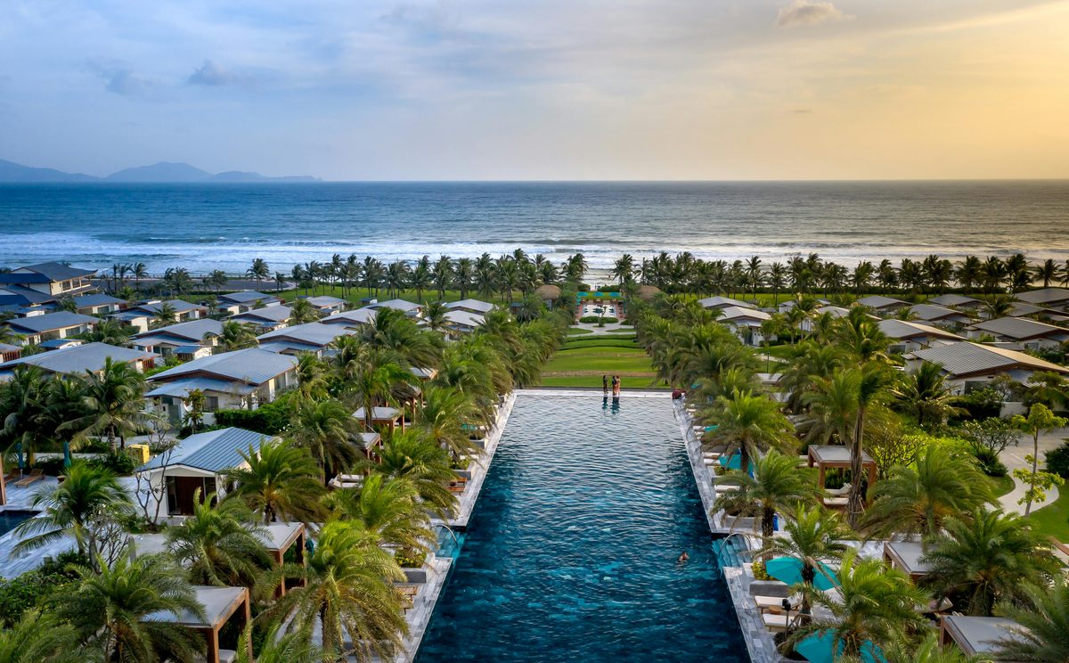 Aerial view of a luxury beachfront resort at sunset with an infinity pool surrounded by palm-shaded villas leading to a sandy beach and ocean horizon
