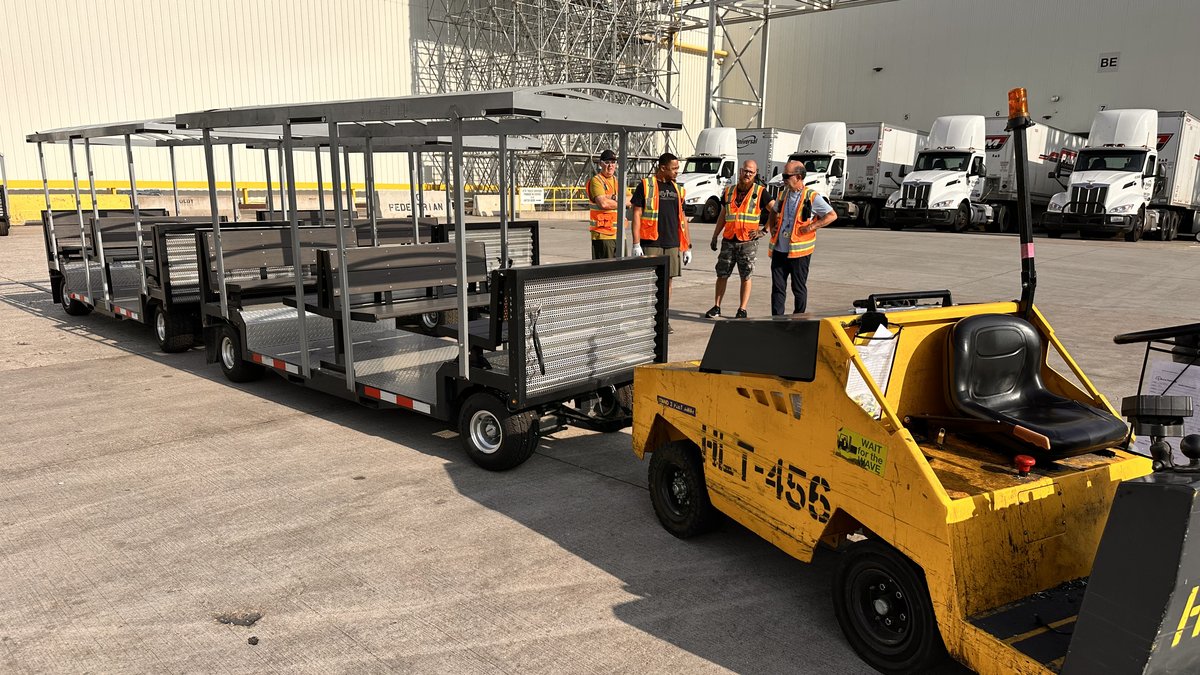 A FlexTram multi-car passenger tram staged in a distribution center yard with workers in safety vests beside it and semi-trailers in the background