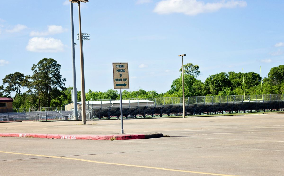 Empty stadium parking lot with worn asphalt, a faded posted-parking sign, and grandstand seating visible in the background under a clear sky