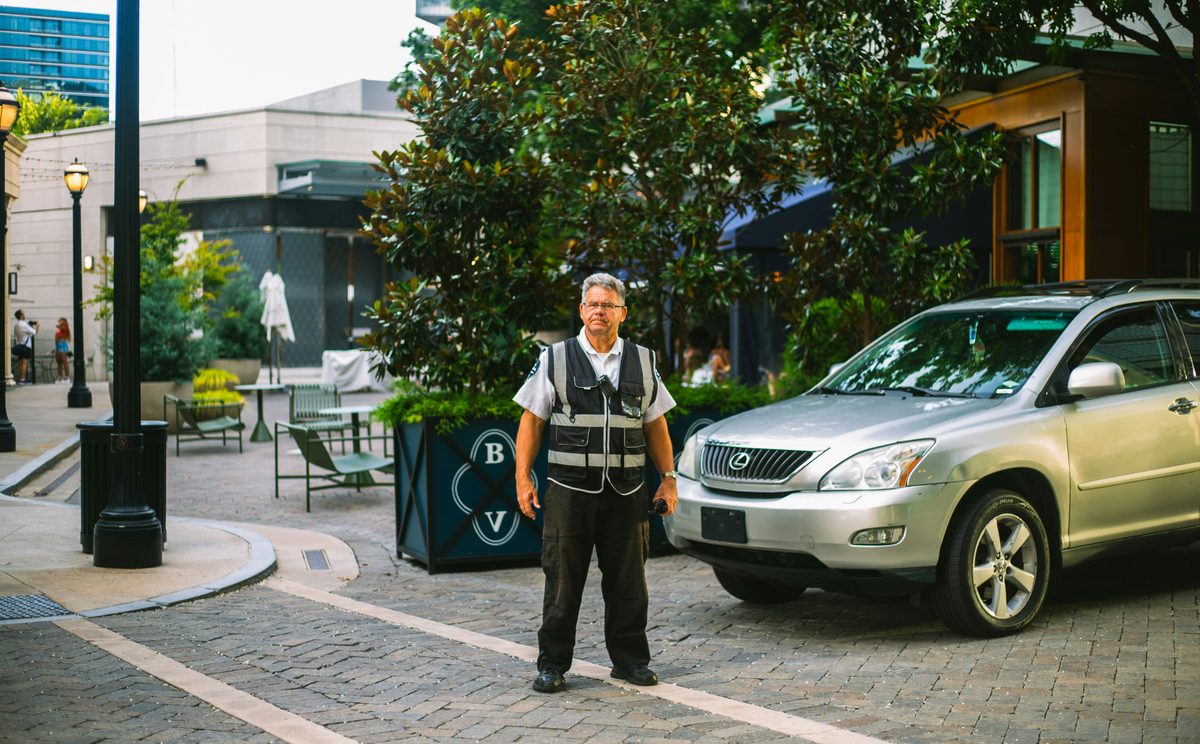 A parking attendant in a reflective safety vest stands in a paved plaza in front of a silver SUV, with a restaurant patio and street lamps behind him