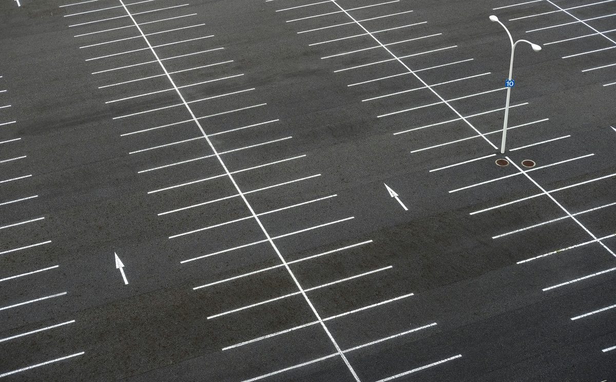 Aerial view of a large empty parking lot showing the angled striped grid pattern, a lamp post, and a '10' zone marker — the surface every modern parking operator manages but where the last 500 yards to the destination remains a gap