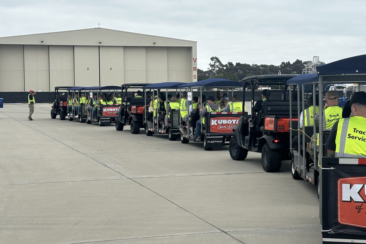 A train of open-air FlexTram trams towed by Kubota utility vehicles carries seated guests in hi-vis vests across a concrete apron past a large aircraft hangar during a facility tour