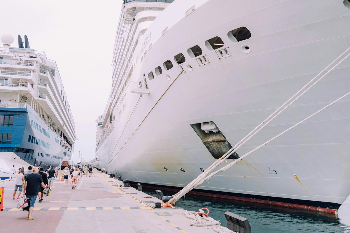 Cruise passengers walking along a pier alongside a docked cruise ship at a port of call, with a second ship visible in the background