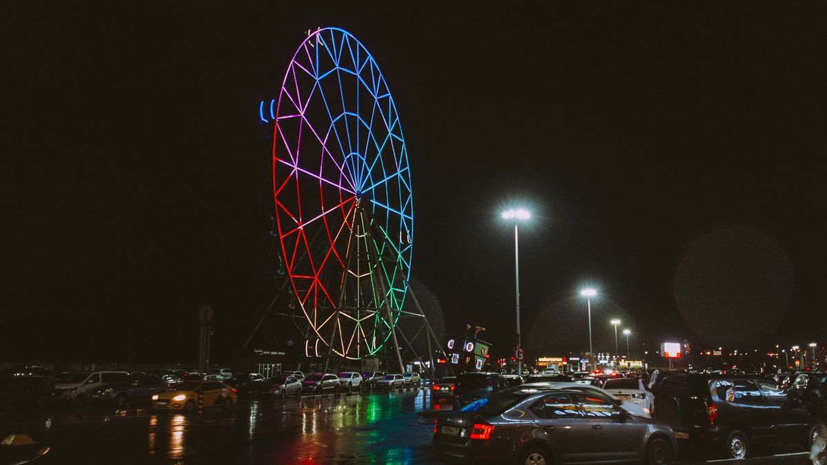 A brightly lit multicolored Ferris wheel towers over a packed, rain-slicked amusement park parking lot full of cars at night