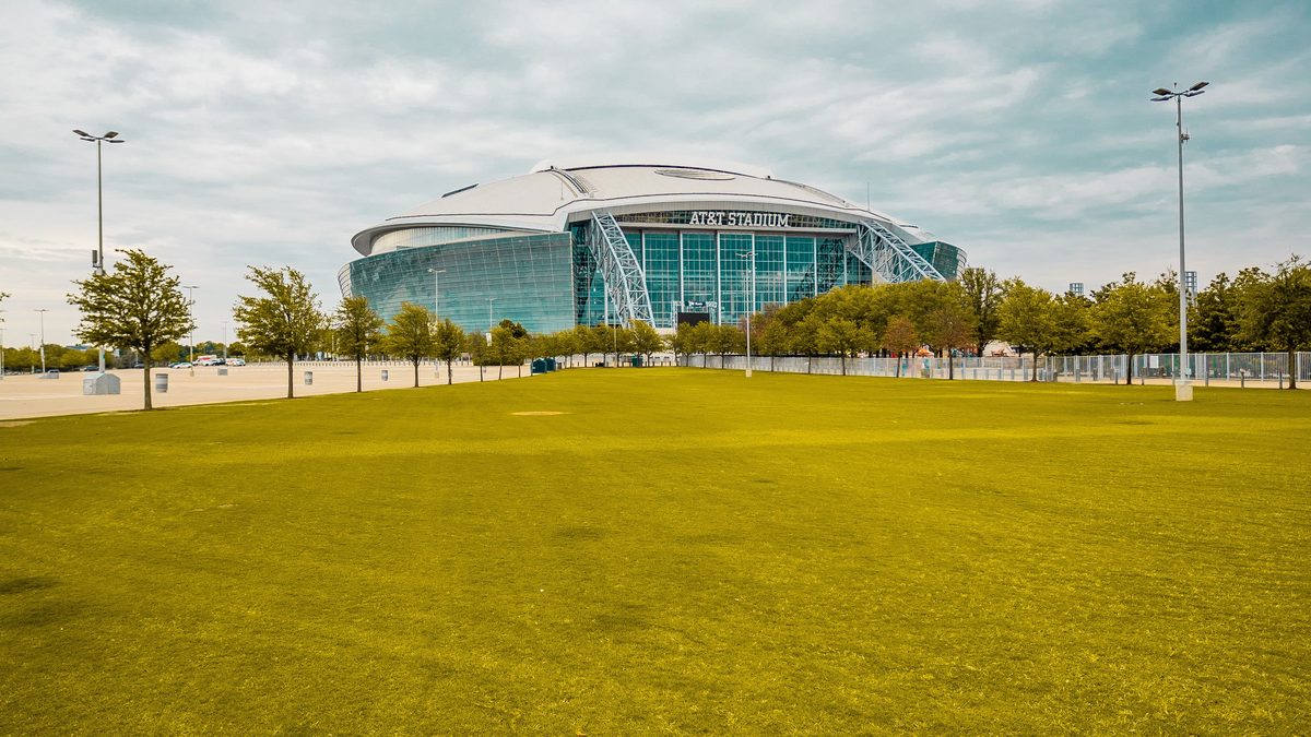 AT&T Stadium in Arlington, Texas, viewed across a wide green lawn under a dramatic cloudy sky