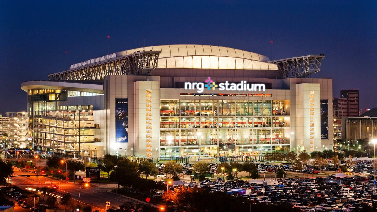 NRG Stadium in Houston lit up at dusk with a full parking lot in the foreground