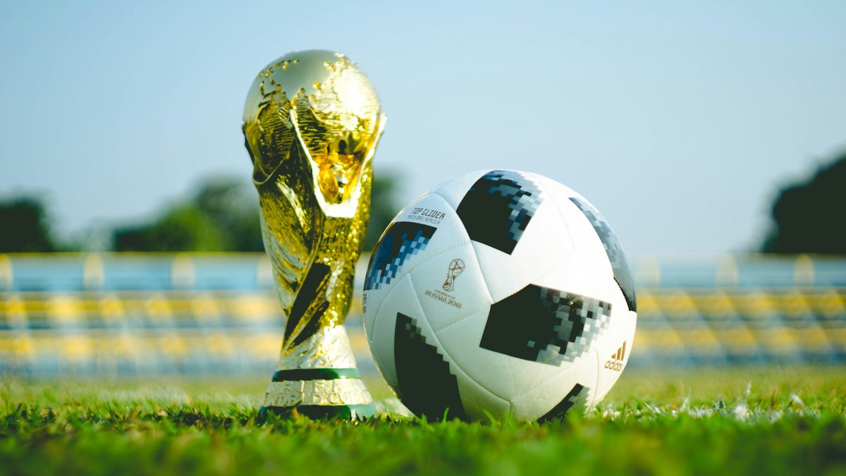 The FIFA World Cup trophy and an official tournament soccer ball resting on a stadium pitch with empty grandstand seating in the background