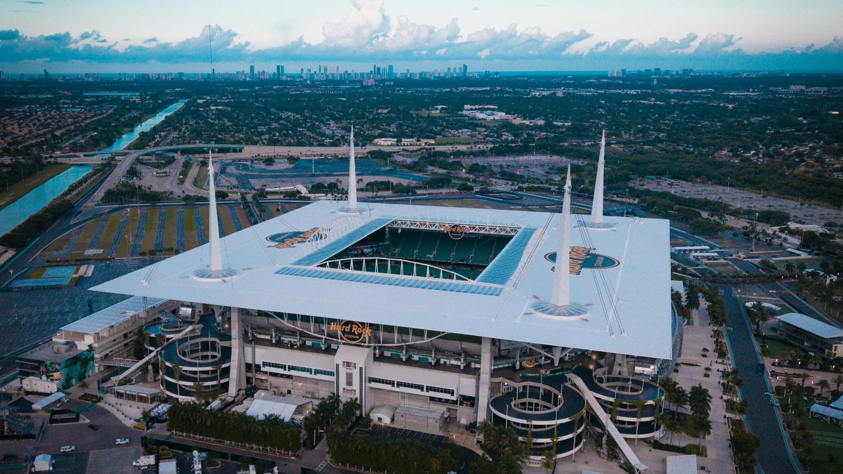 Aerial view of Hard Rock Stadium in Miami Gardens at dusk, surrounded by vast parking lots with the Miami skyline on the horizon