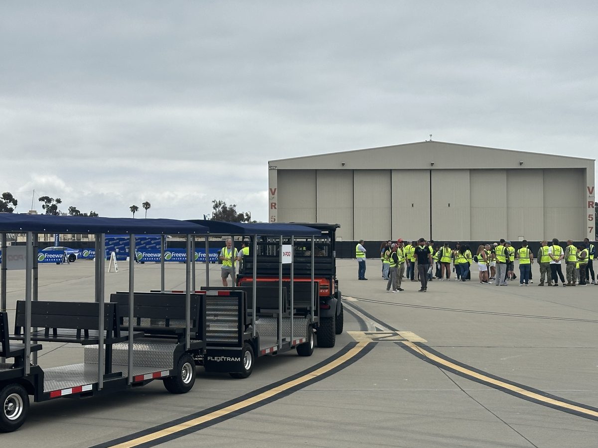 A FlexTram crew shuttle staged on an airfield apron beside a hangar while staffing workers in safety vests gather for transport
