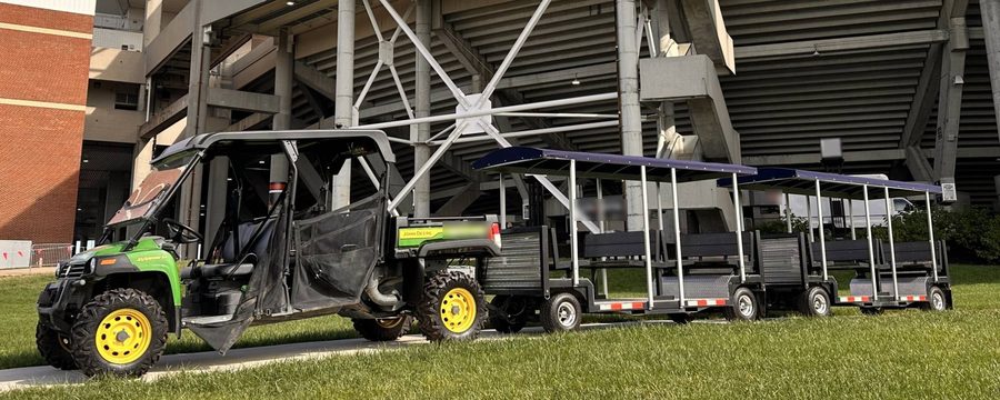 FlexTram tram cars towed by a utility vehicle on the lawn outside a college football stadium