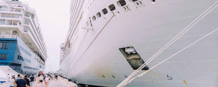 Cruise passengers walking along a pier alongside a docked cruise ship at a port of call, with a second ship visible in the background